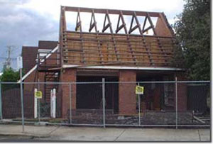 Photo of a house which is partly demolished. Brick piers are standing on the ground level, and the timber roof structure is visible and incomplete.