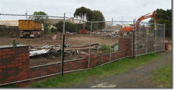 Photo of a fenced site with an excavator clearing the debris. There is also a truck at the site. 